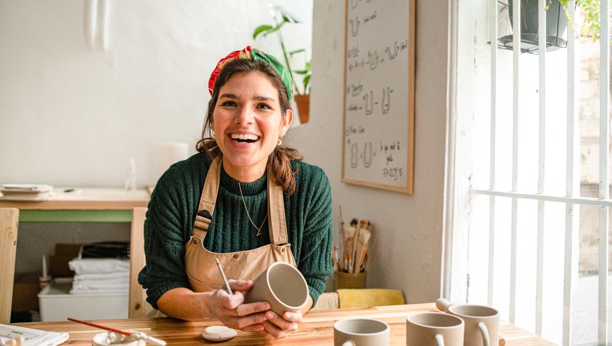 A happy female ceramicist smiles at the camera while working on clay cups at her art studio
