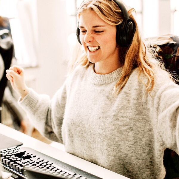 A young woman wearing headphones making a triumphant gesture behind the computer at the office desk