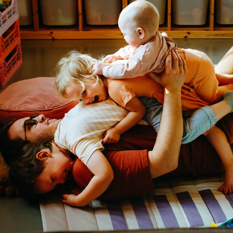 Three young children playfully lay on top of their father on the floor at home