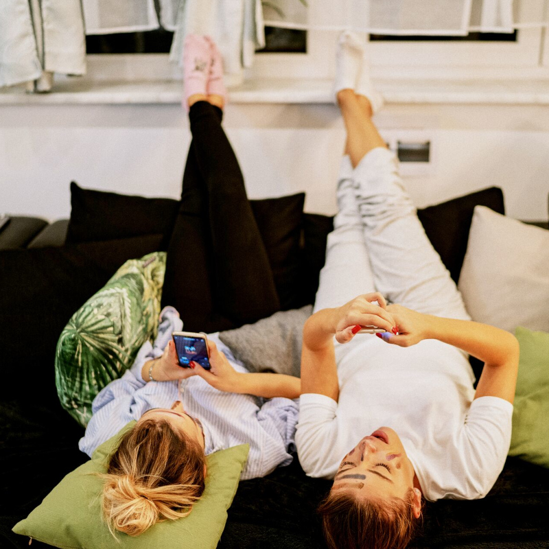 Two female friends lay on the couch at home with their legs up while scrolling on their phones