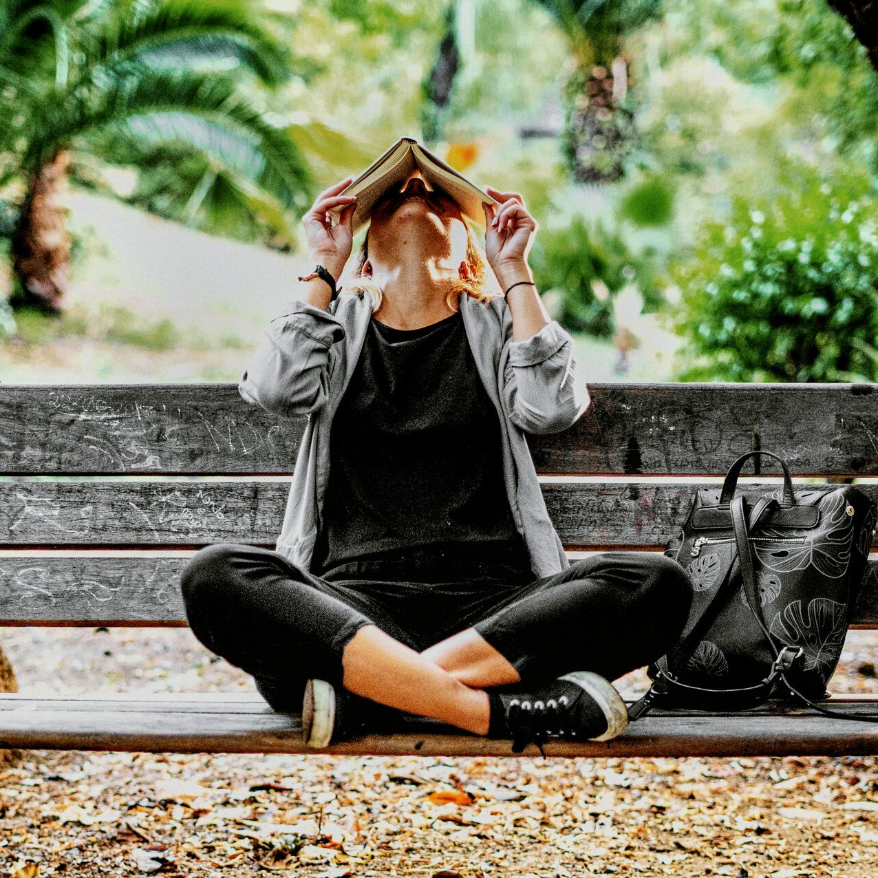 Woman sitting cross legged on a bench in the park covering her face with a book 800